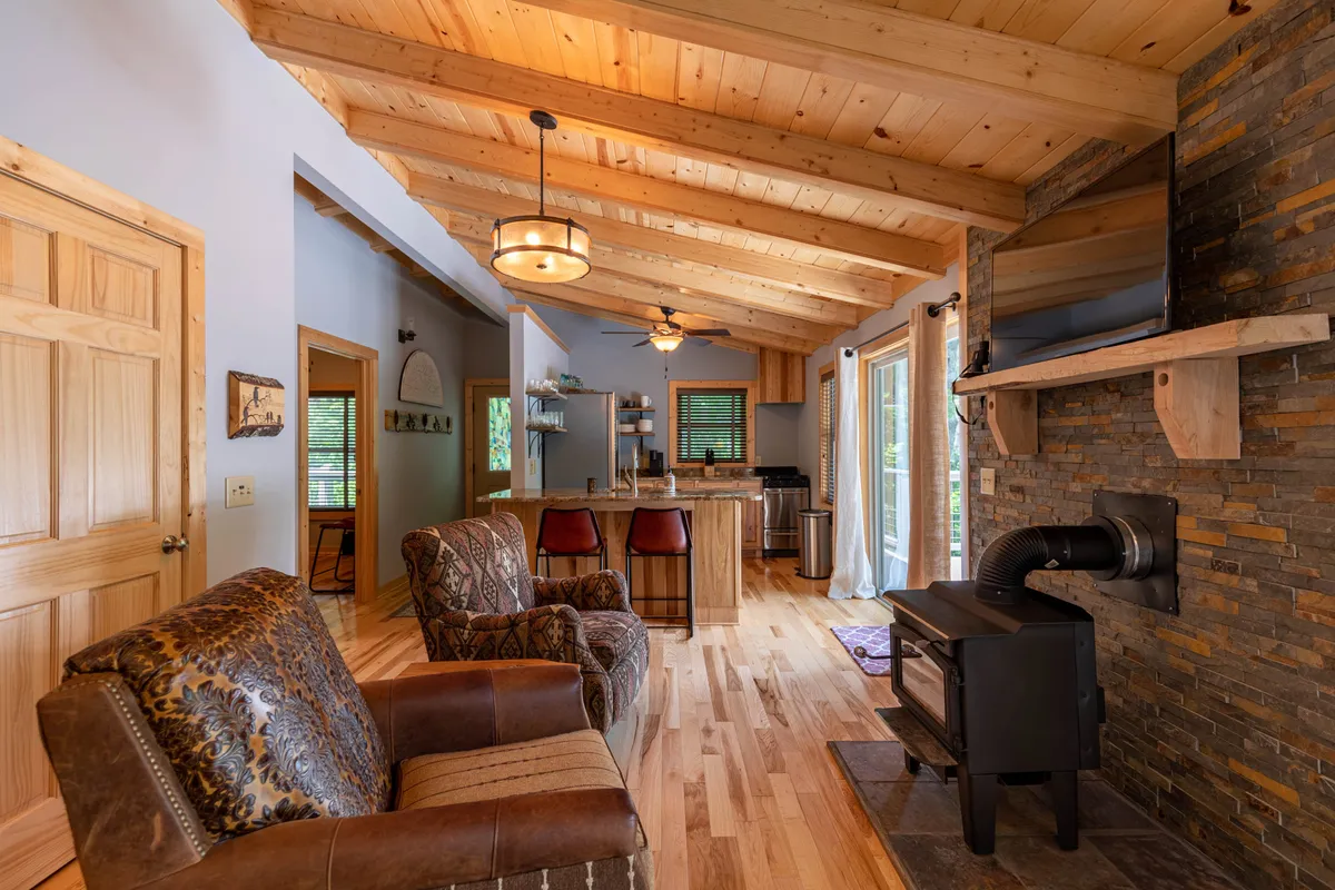 A cozy view from the living area shows wooden ceiling beams, a stone fireplace, and bar stools lining the kitchen counter, hinting at the seamless connection between conversation and culinary activity. Comfortable seating invites relaxation, making this a natural transition space between socializing and dining.