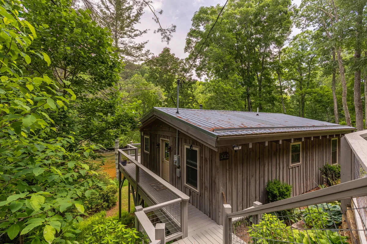 An inviting exterior of a wooden cabin nestled in greenery, highlighting a spacious deck area with seating. The scene suggests opportunities to relax on the deck, enjoy sunrises, and watch for wildlife in the comfort of the outdoors.