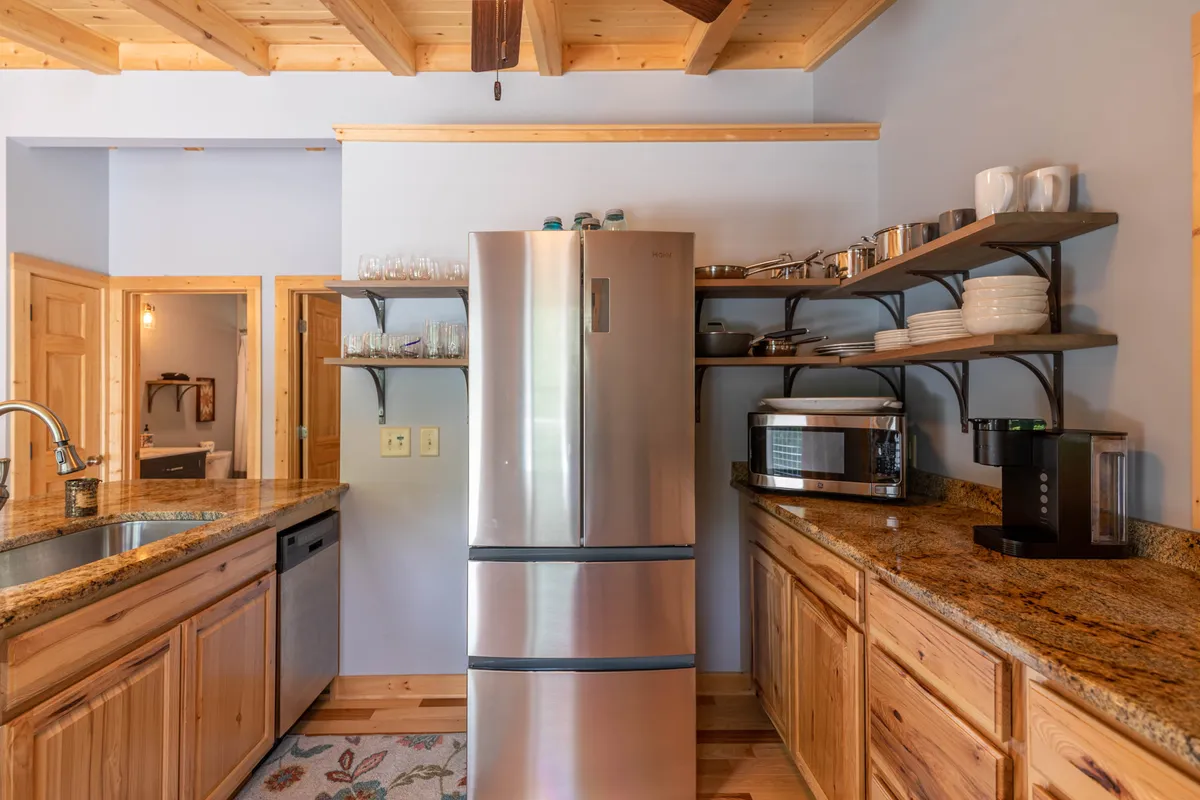 This kitchen scene highlights stainless steel appliances, including a large refrigerator, and broad granite countertops. Open shelving with neatly arranged dishes and natural wooden cabinetry create a warm and functional atmosphere, ideal for cooking and entertaining guests.