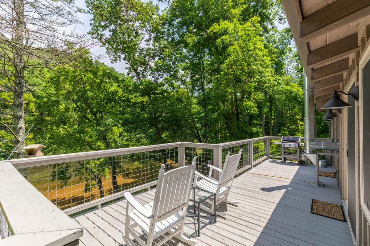A cozy outdoor deck setup with deck chairs and a grill, bordered by a rustic wooden railing. The area is surrounded by trees and natural foliage, creating a secluded and peaceful retreat. The setup suggests both relaxation and opportunities for outdoor dining or gatherings while immersed in nature.