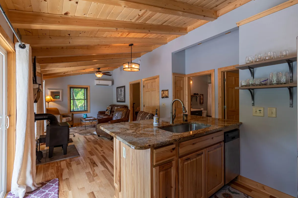This image offers a wider view of the open living area, showcasing the seamless transition between the kitchen and living room. Distinctive wooden beams overhead and a cozy seating area are visible, along with a kitchen island perfect for informal meals or gathering with loved ones. Soft natural light enhances the overall warmth of the space.