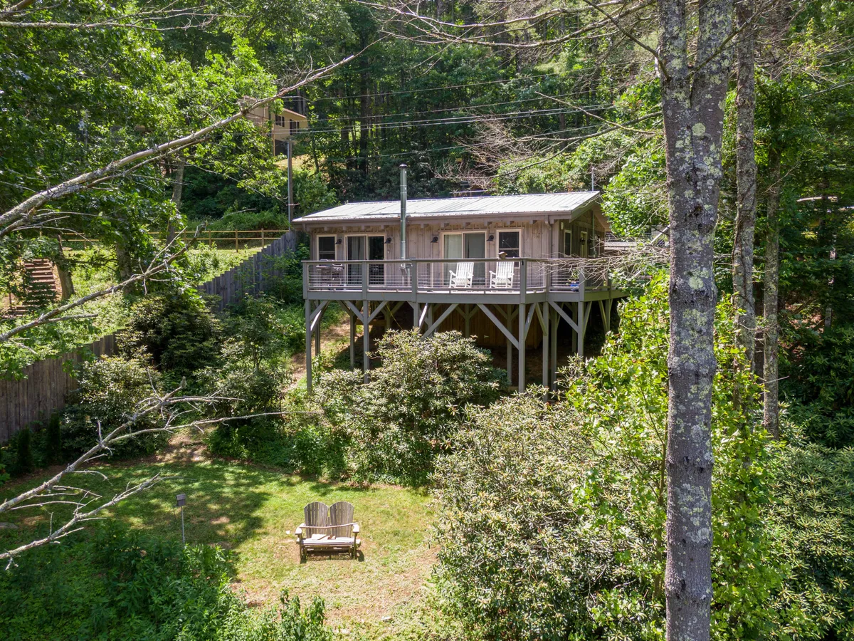 The image shows an elevated house surrounded by lush greenery, featuring an inviting outdoor seating area on the deck. The natural setting showcases open grassy areas and hints of nearby nature trails, creating a tranquil environment perfect for outdoor activities with pets. The deck provides a comfortable spot to relax and take in the scenic views, making it ideal for watching sunsets or supervising pets as they play.
