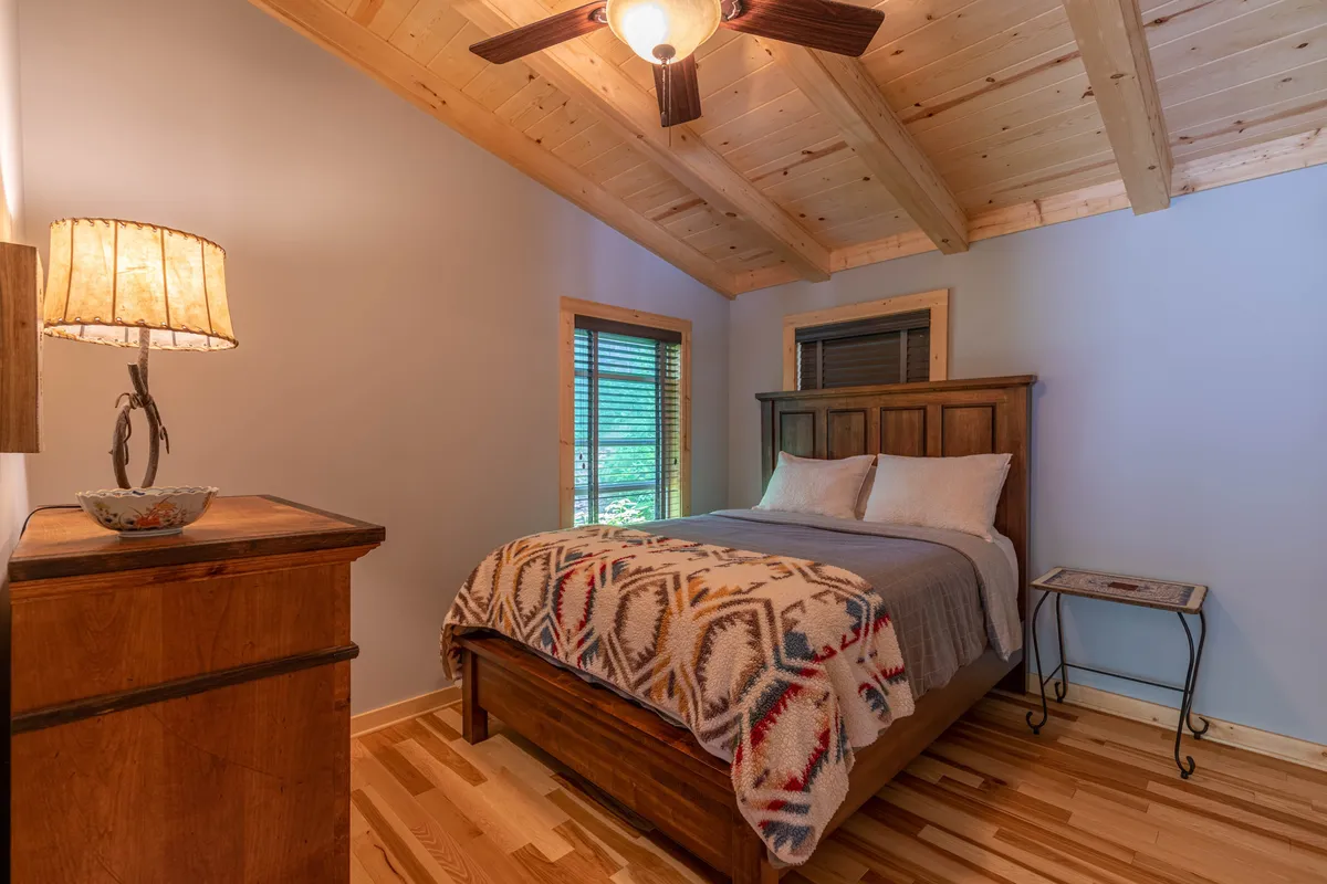 A cozy bedroom with a bed covered in a patterned blanket, warm wooden furniture, and a decorative lamp. Sunlight gently filters in through the blinds, creating a serene, restful atmosphere ideal for relaxation and sleep.