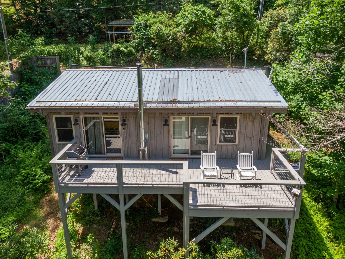 The image showcases a charming wooden cabin with a spacious deck featuring inviting chairs, all nestled amidst lush greenery. The deck overlooks the tranquil South Fork New River, providing an idyllic spot to relax and soak in peaceful nature views. The scene is set during golden hour, hinting at beautiful sunset vistas and opportunities for morning coffee or evening stargazing.