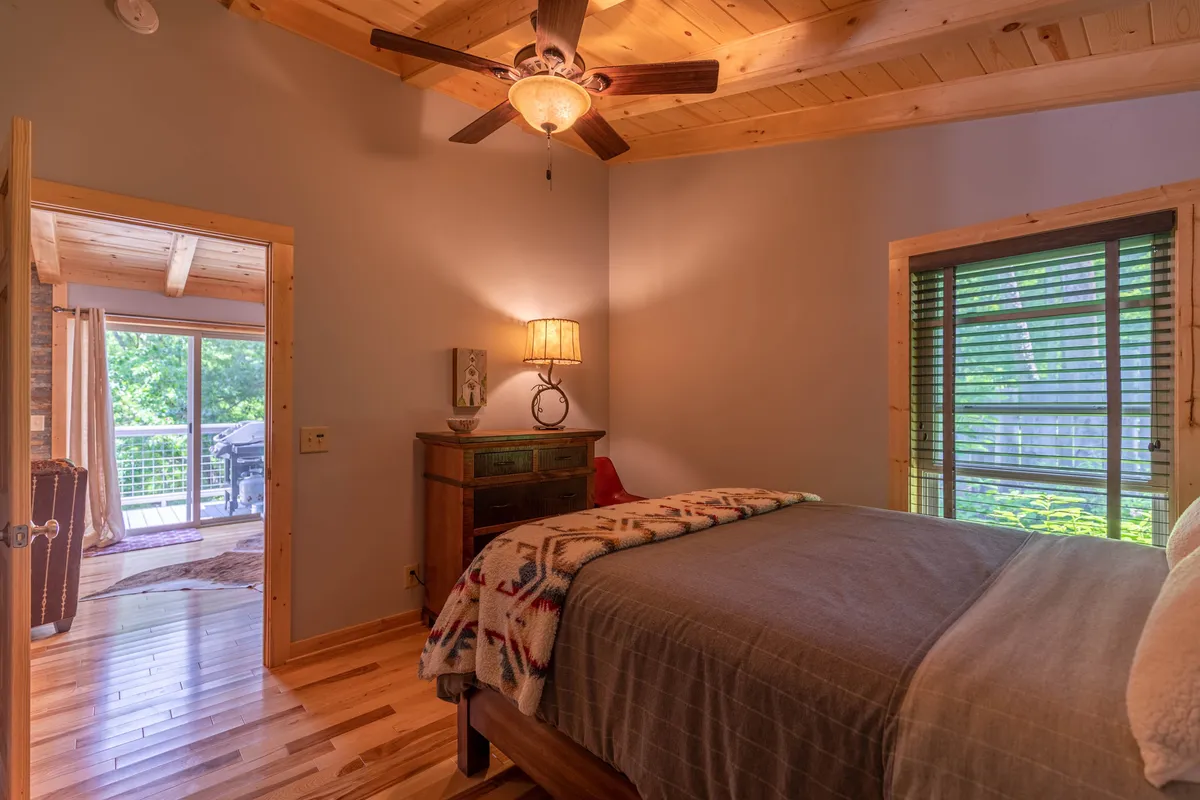 A calm and serene bedroom scene featuring a comfortable bed, wooden furniture, a bedside lamp, and a window letting in soft natural light. The inviting setup suggests relaxation and the enjoyment of scenic views from the window.