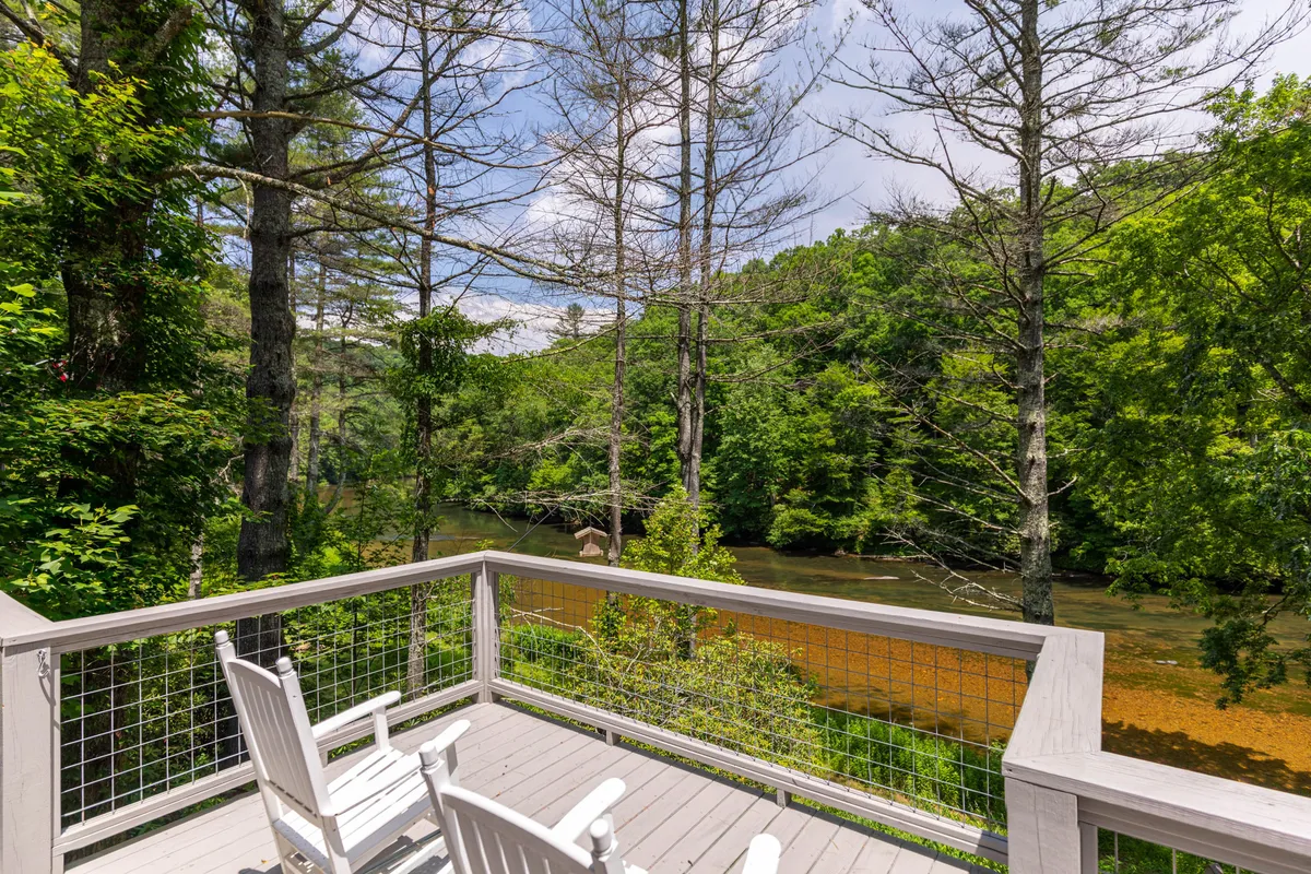 This image features a spacious wooden deck overlooking a scenic river, bordered by lush greenery. On the deck, comfortable rocking chairs are arranged to invite relaxation and enjoying the tranquil views. The peaceful mood is enhanced by the warm, natural light and the clear sightlines to the river, evoking a sense of calm and outdoor serenity.