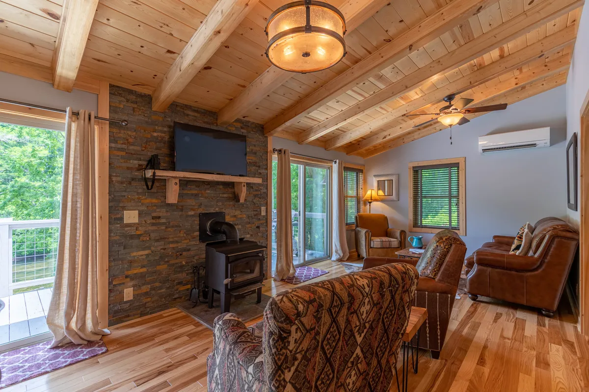 This image portrays a warmly lit living room featuring a striking stone fireplace, exposed wooden beams, inviting leather seating, and large windows that allow natural light to pour into the space. The scene is arranged to encourage conversation and relaxation, and the fireplace speaks to cozy gatherings, matching the section’s emphasis on warmth and togetherness.